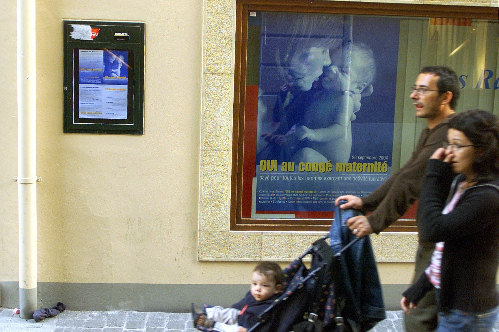 En 2004, un couple avec enfant passe devant une affiche de la campagne pour le oui au congé maternité à Neuchâtel.