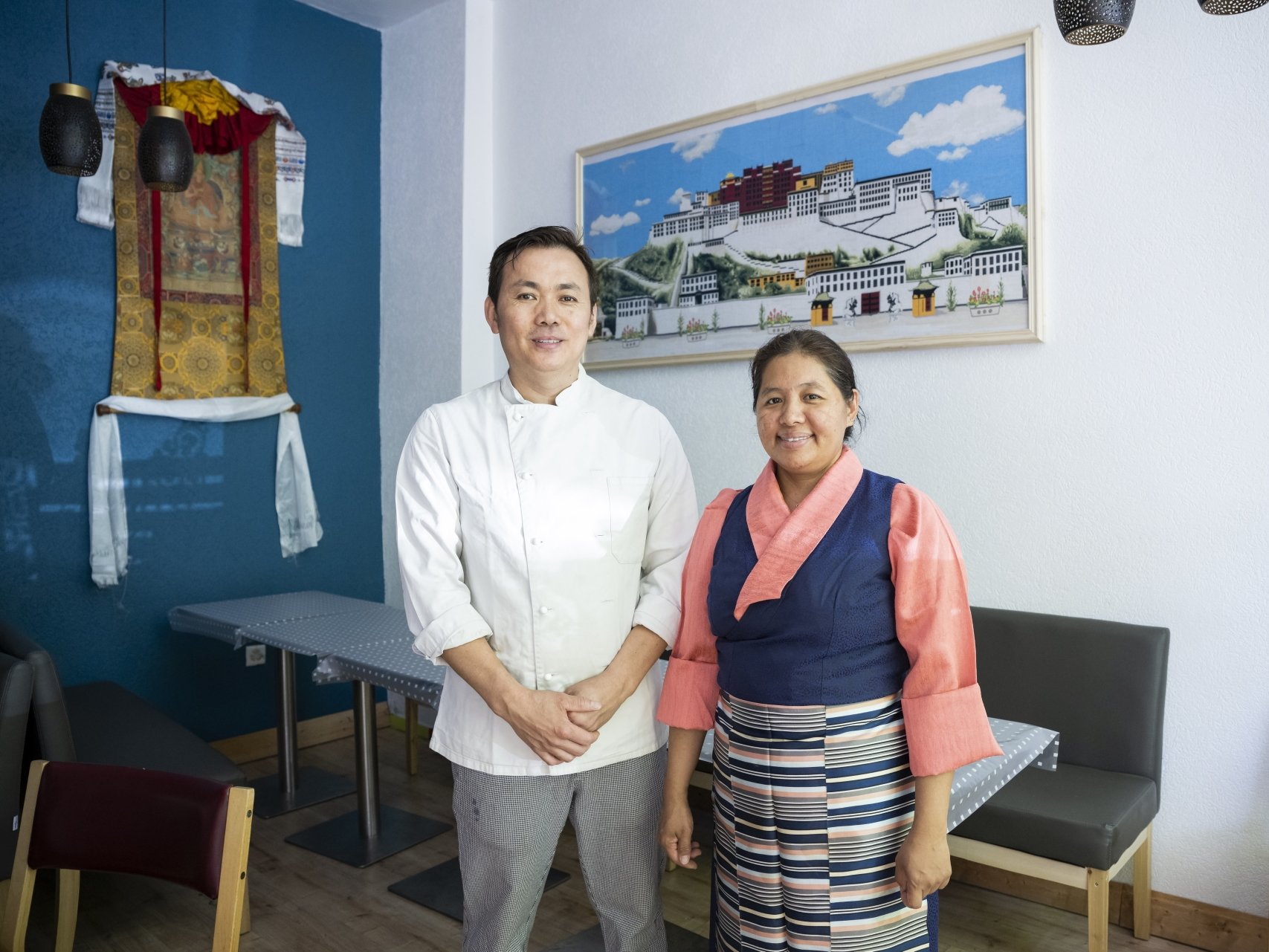 Yonten et Dawa Tratsang - elle porte le costume traditionnel - posent dans la petite salle de leur restaurant. Derrière eux, une broderie représentant Lhassa, capitale du Tibet.
