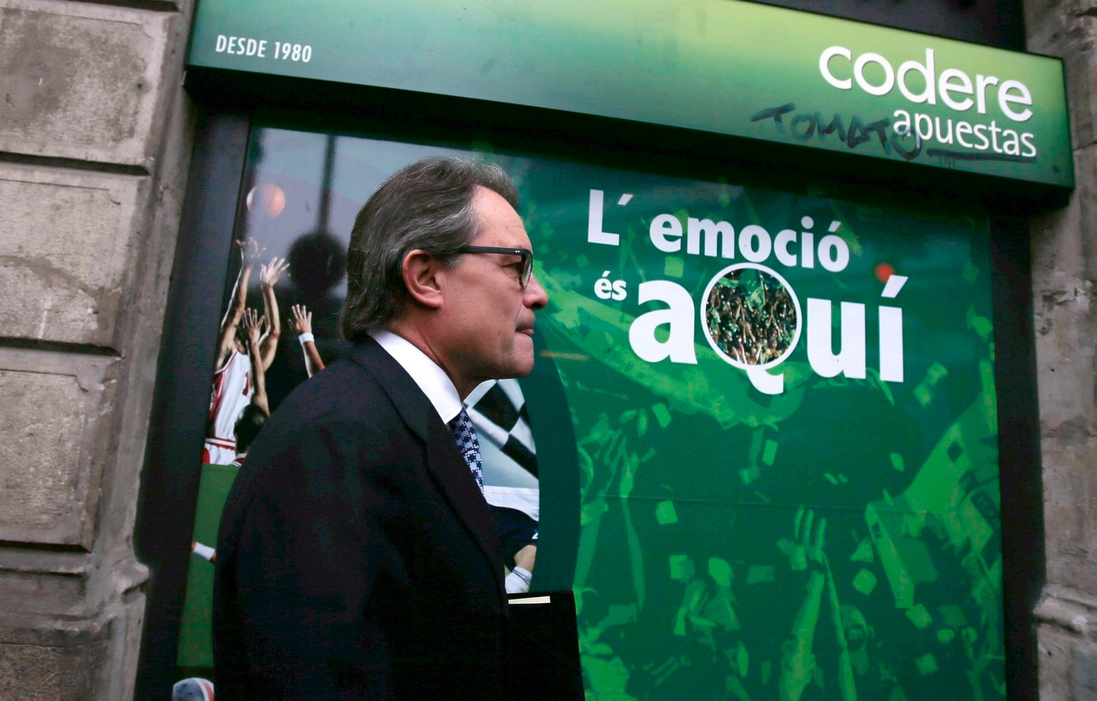 Acting regional President Artur Mas walks past a betting shop with a banner reading in Catalan: "The excitement is here" in Barcelona, Spain, Monday, Jan. 4, 2016. Catalonia's two main pro-independence parties will meet to discuss how to salvage their secessionist drive after a key far-left party refused to back the re-election of incumbent regional leader Artur Mas. Mas' ruling conservative Convergence party and the Republican Left of Catalonia group joined forces under the "Together for Yes" alliance to win 62 seats in the 135-seat regional parliament last September. But they needed the support of the far-left CUP's 10 seats to secure Mas a workable majority and further the independence push. (AP Photo/Manu Fernandez) Spain Catalonia Independence