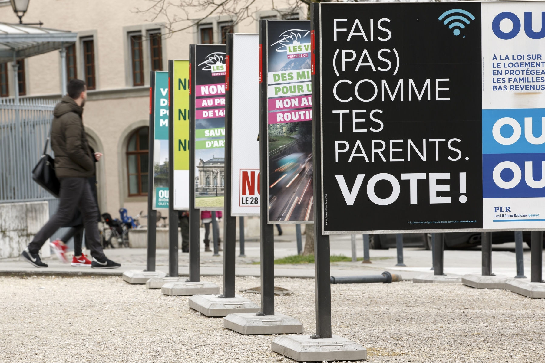 Des personnes passent vers l'affiche "Fais (pas) comme des parents. Vote!" du Conseil d'Etat genevois, et d'autres affiches concernant les votations du 28 fevrier, ce mercredi 24 fevrier 2016 a Geneve. Le Conseil d'Etat genevois a fait placarder des affiches pour incite les jeunes a voter. (KEYSTONE/Salvatore Di Nolfi)