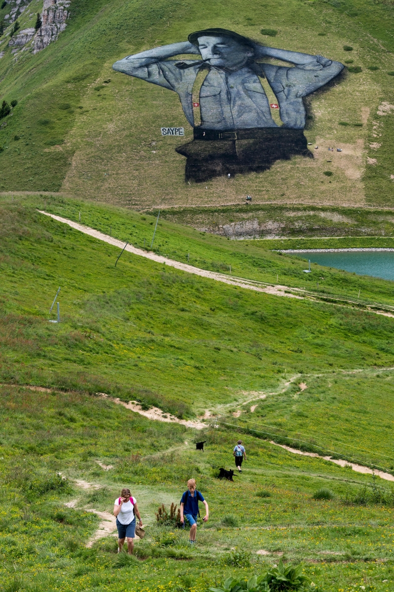 Des personnes marchent devant une fresque sur herbe representant un berger suisse de 10'000Äâm2 en peinture biodegradable faite par l'artiste francais Saype et visible a plus de 2000 metres d'altitude ce jeudi 4 aout 2016 a Leysin dans les alpes vaudoises. (KEYSTONE/Jean-Christophe Bott) SUISSE LAND ART SAYPE