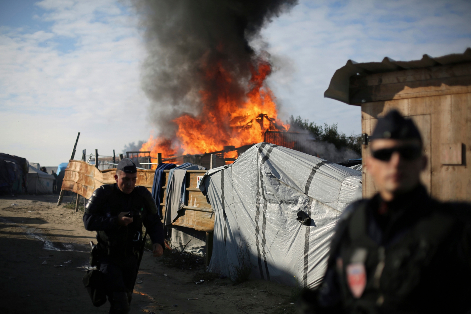 Riot police stand guard in front of a burning wooden house in the makeshift migrant camp known as "the jungle" near Calais, northern France, Tuesday, Oct. 25, 2016. Crews have started dismantling the migrant camp in France after the process to clear the camp began in earnest on Monday. (AP Photo/Thibault Camus) France Migrants