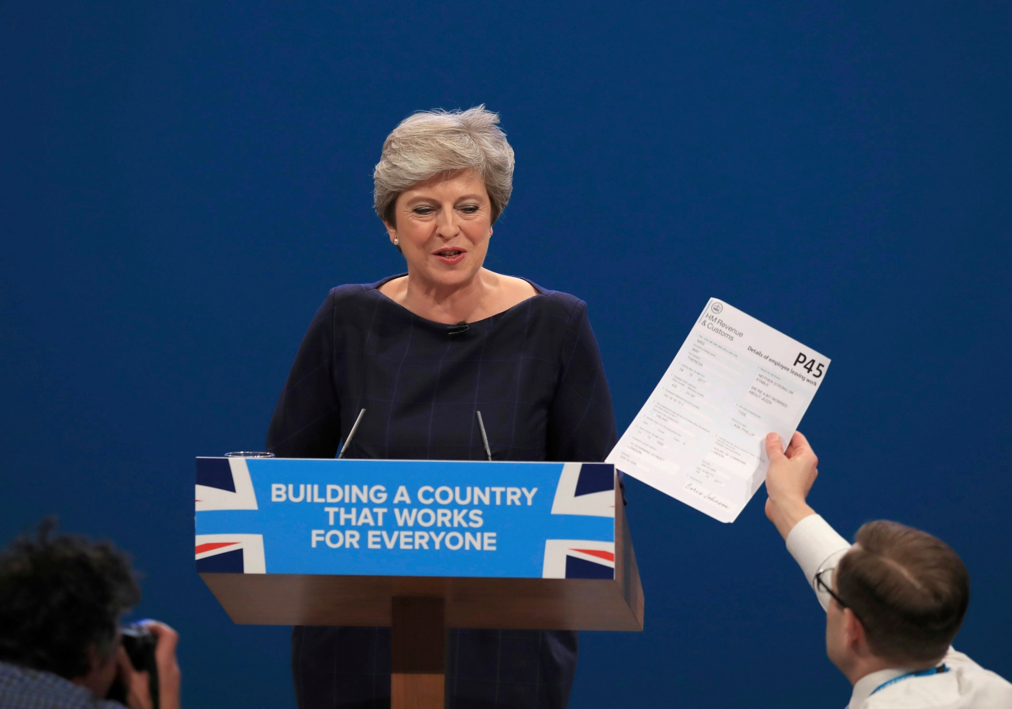 Comedian Simon Brodkin, also known as Lee Nelson confronts British Prime Minister Theresa May during her keynote speech, during the Conservative Party Conference at the Manchester Central Convention Complex in Manchester, England, Wednesday Oct. 4, 2017. (Peter Byrne/PA via AP) APTOPIX Britain Conservatives