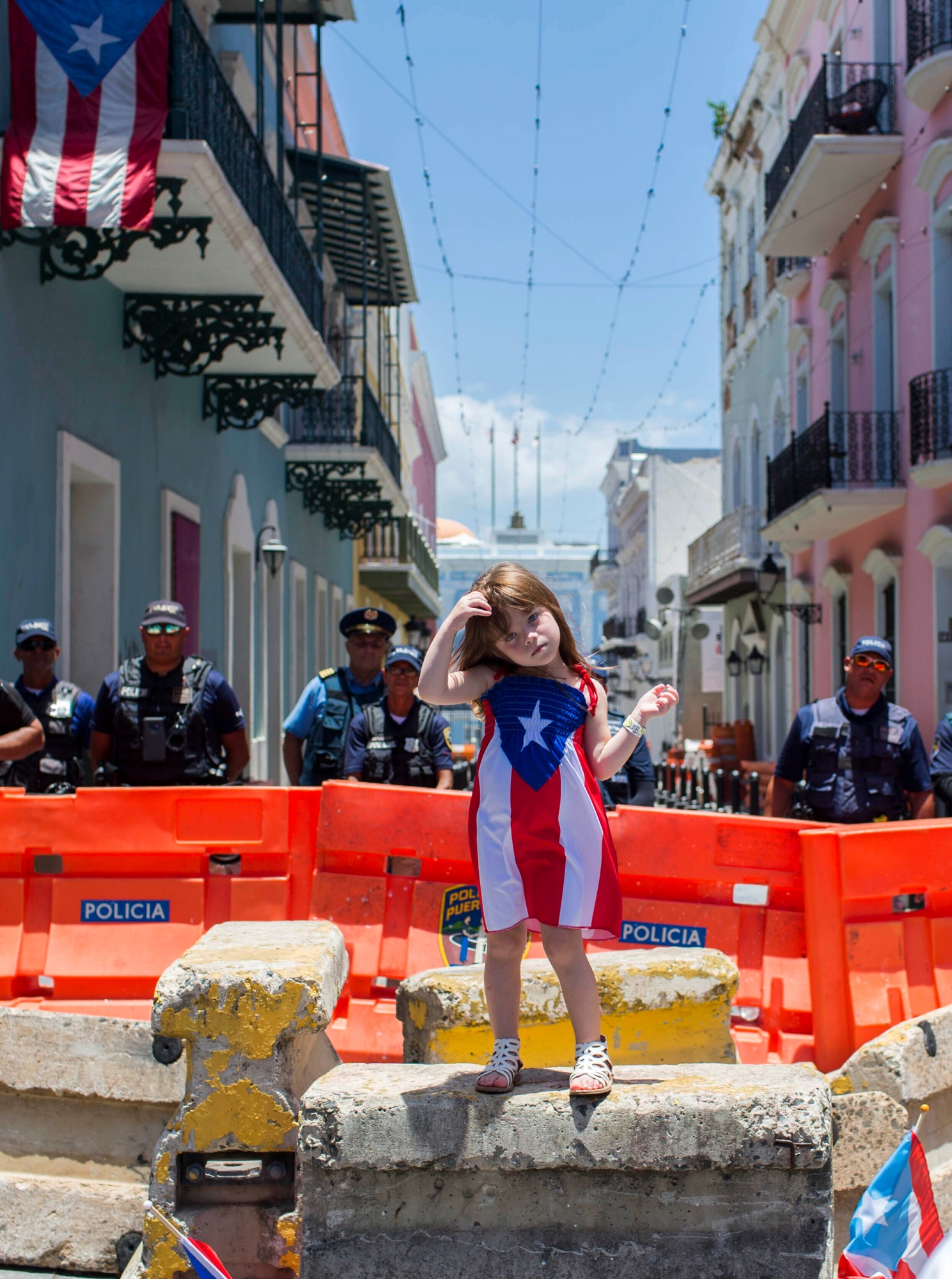 A girl wearing a dress featuring the Puerto Rican flag stands by police blocking the road leading to the La Fortaleza governors mansion in San Juan, Puerto Rico, Thursday, July 18, 2019. Protesters are demanding Gov. Ricardo Rossello resign after the leak of online chats that show him making misogynistic slurs and mocking his constituents. (AP Photo/Dennis M. Rivera Pichardo) PUERTO RICO PROTESTS